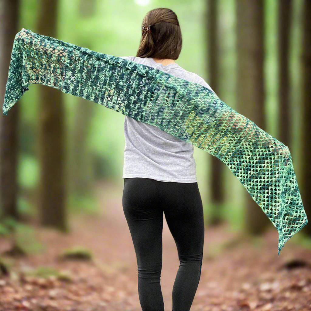 model holding out ariel shawl in variegated green colorway with greenery in the background.