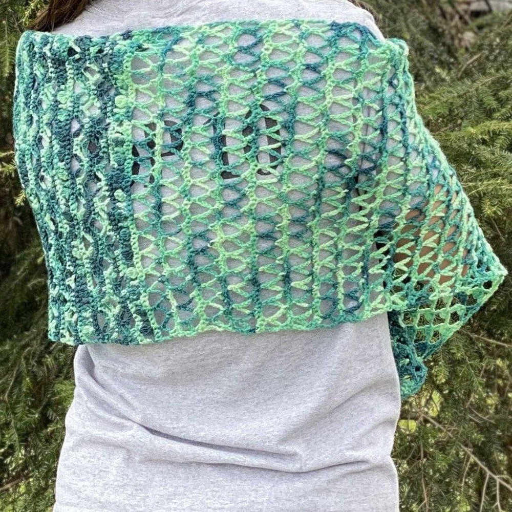 model wearing ariel shawl in variegated green colorway with greenery in the background.