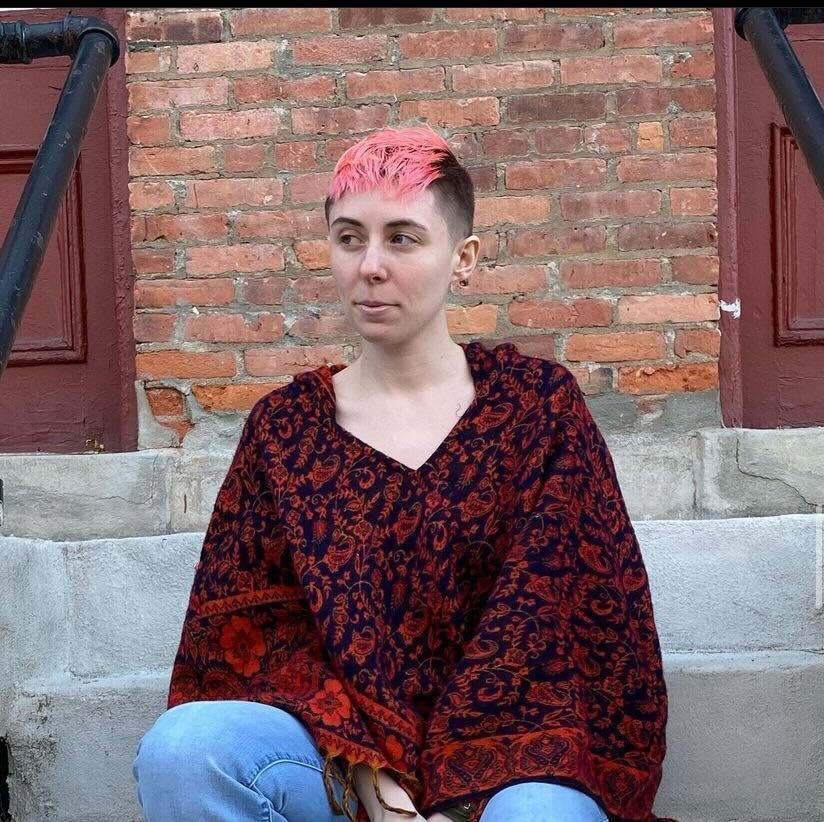 Profile picture of the author, Kate Curry, wearing a dark red Nanda Poncho sitting on concrete stairs in front of brick wall.
