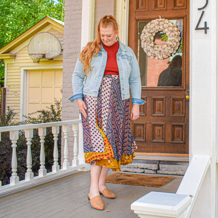 Woman on a porch in red top, patterned skirt, and denim jacket, with wooden door and wreath behind.