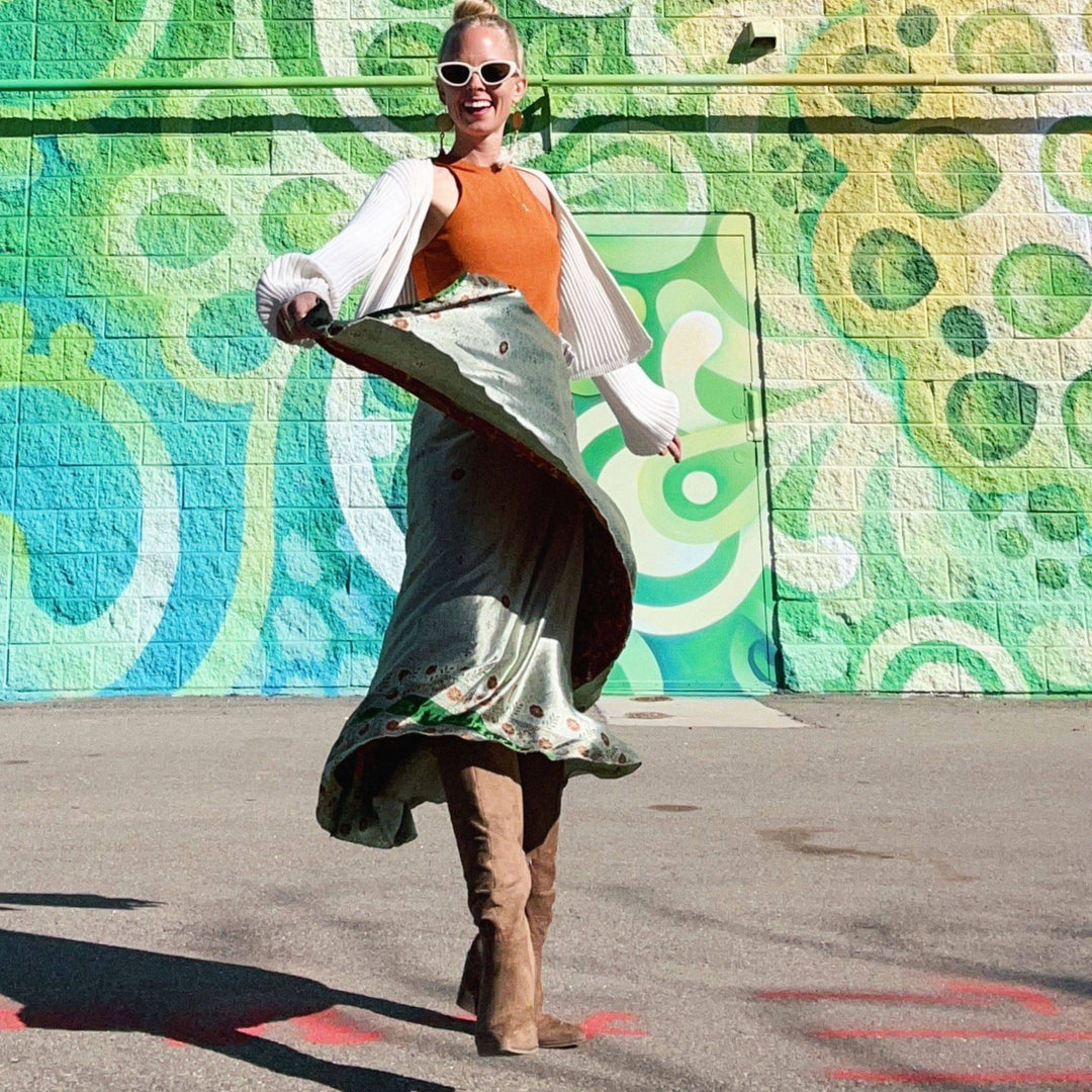 A person wearing a colorful outfit is joyfully spinning in front of a vibrant, abstract mural. They have on a long, flowing skirt, an orange top, a white cardigan, and knee-high boots. The mural features green and blue circular patterns. The person is also wearing white sunglasses and smiling.